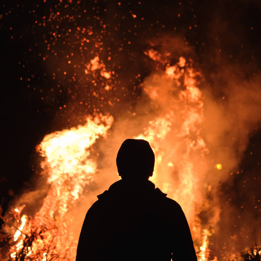 Person in a jacket and beanie standing in front of a raging fire at night.